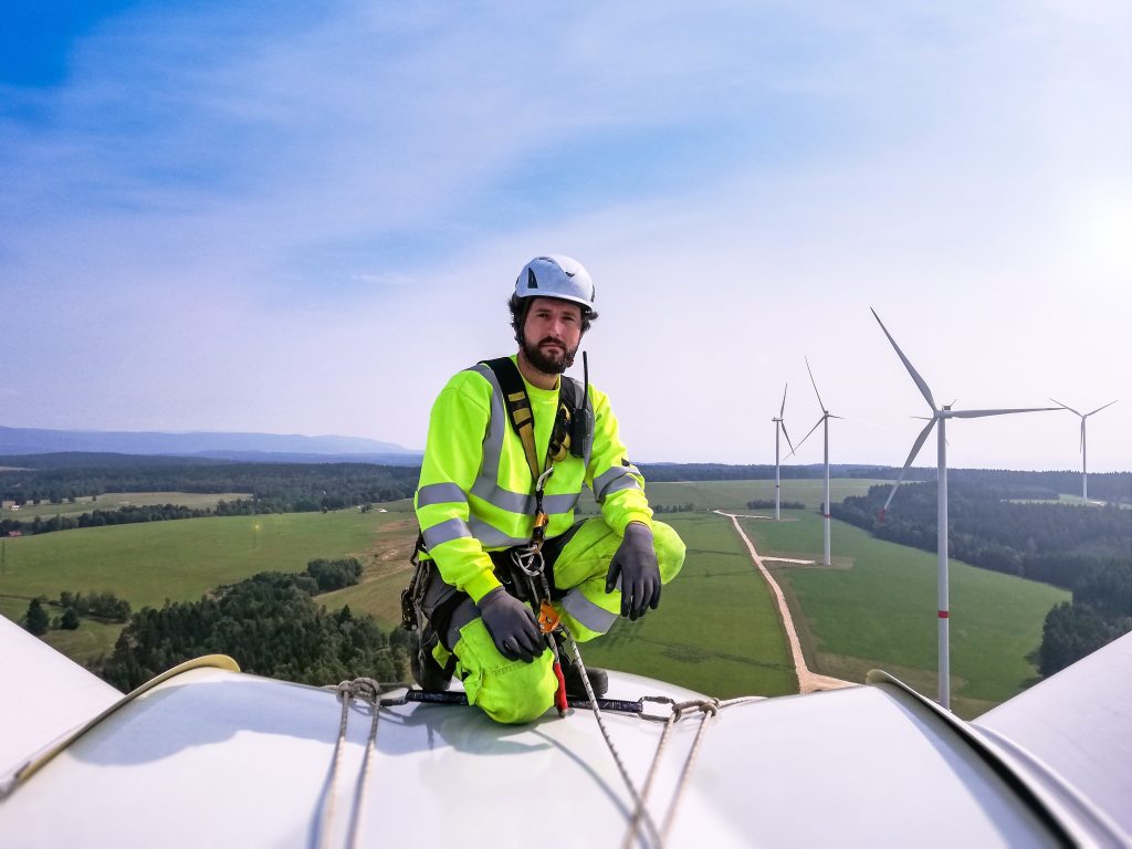 Industrial climber kneeling on a large onshore wind turbine, preparing for blade inspection with a wind farm in the background.