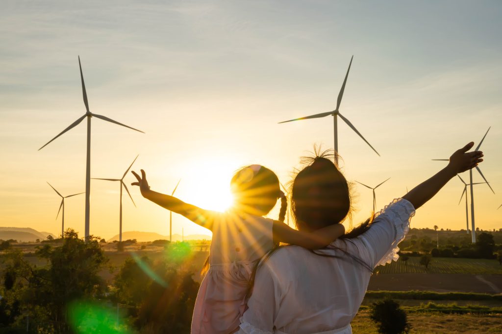 Woman holding a child standing in front of wind turbines with sunlight shining through.