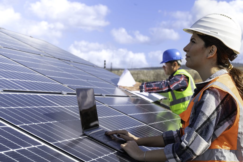 Woman working on a laptop atop a solar panel, with a man holding papers in the background.